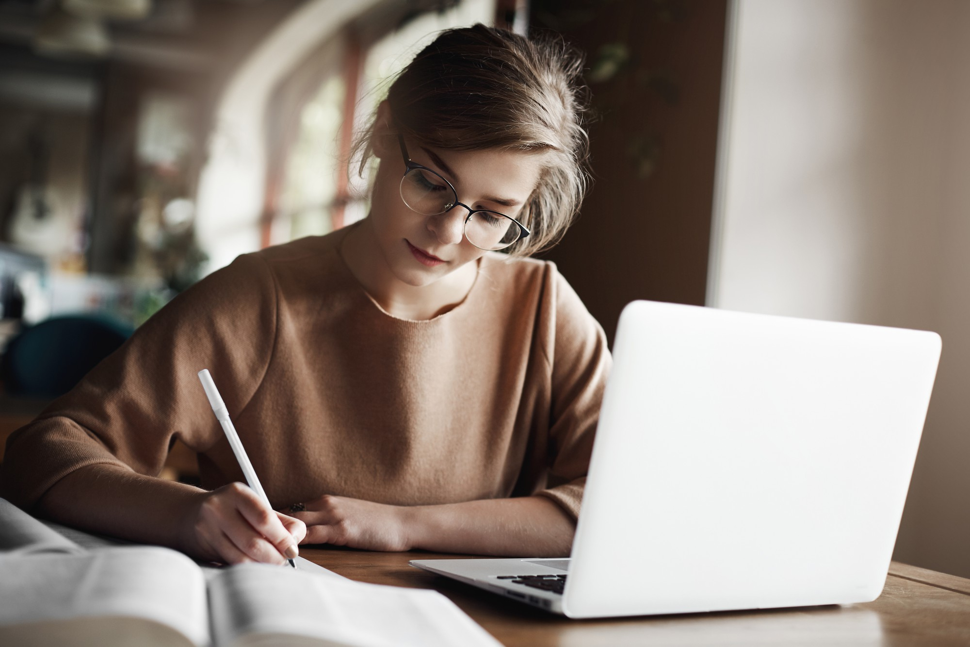 hardworking focused woman in trendy glasses concentrating on writing essay sitting in cozy cafe near laptop working and making notes carefully